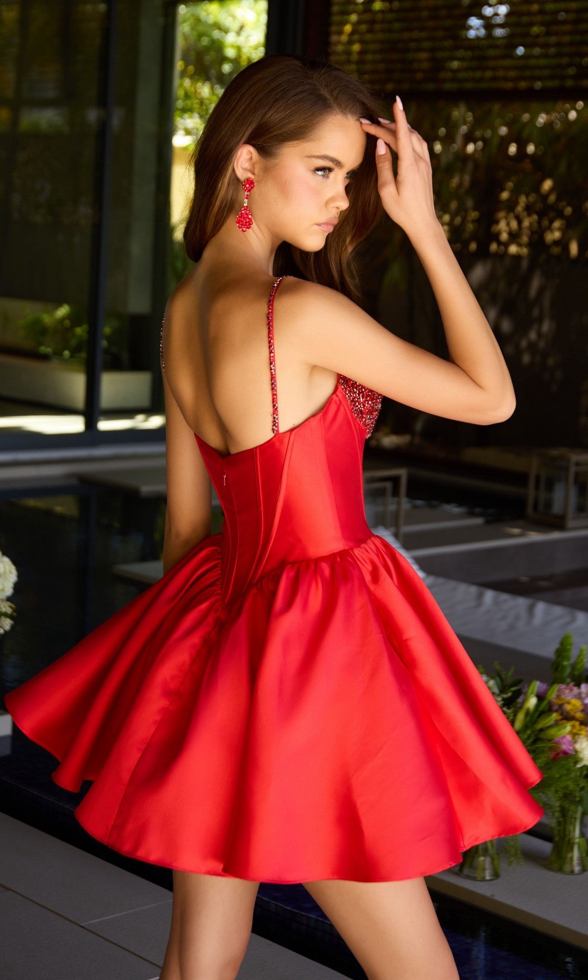 Wearing the Ava Presley 42380, a sleeveless, backless red satin short homecoming dress with a flared skirt, a woman stands poolside, touching her hair and glancing over her shoulder.